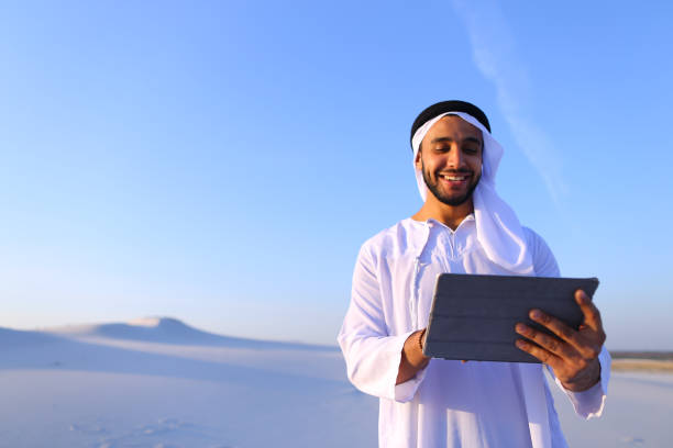 A man in traditional attire smiles while using a tablet in a sandy desert, representing RAKEZ Free Zone business benefits.